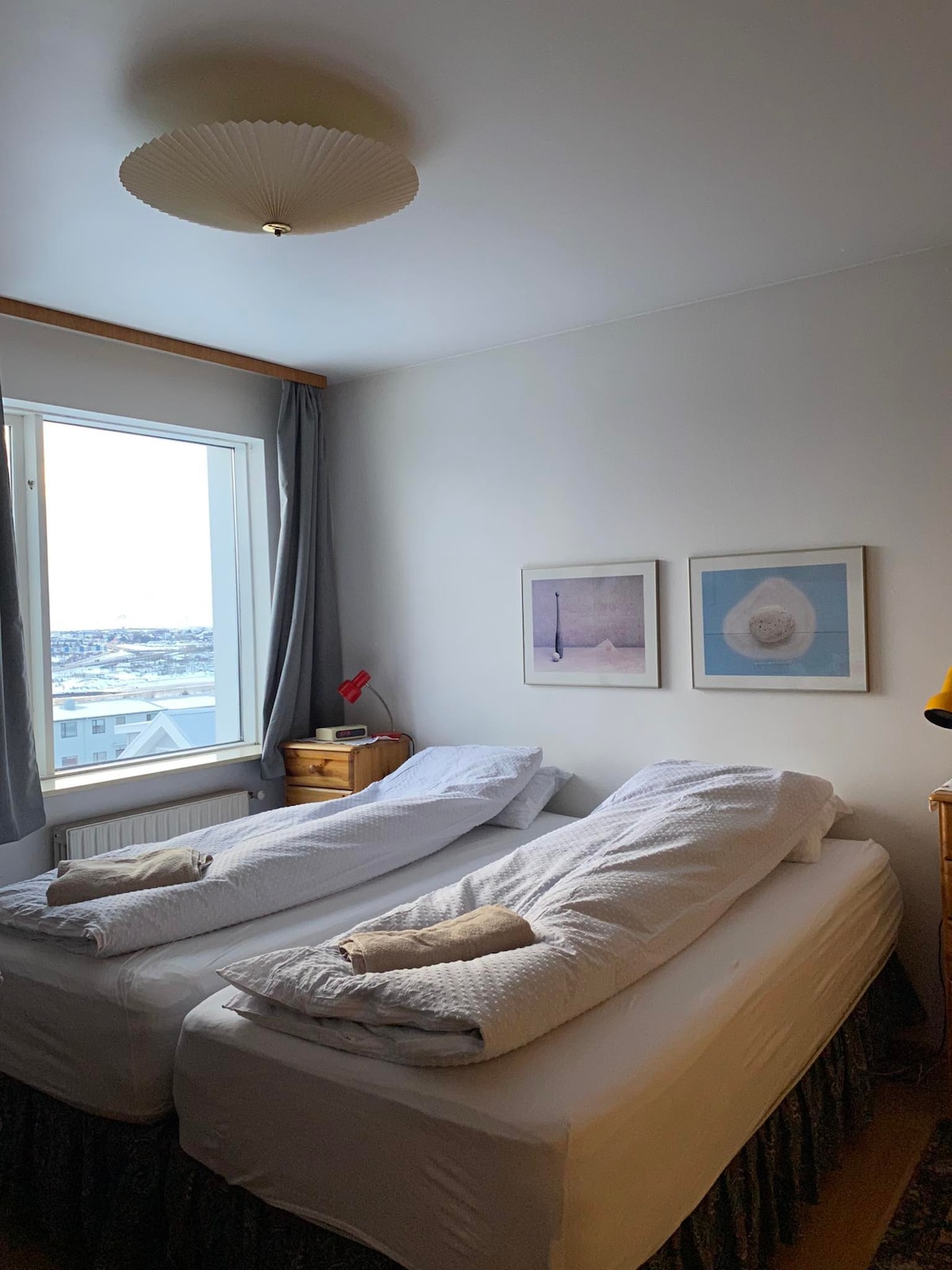 A calm bedroom features two spacious beds with white linens and decorative pillows. Light streams in through a large window, revealing views of the Reykjanes peninsula. The room is complemented by framed artwork and a wooden nightstand with a small lamp.