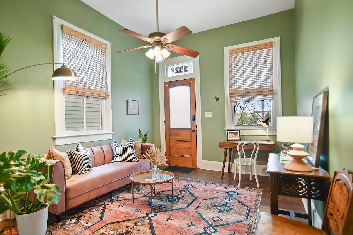 A cozy living area features a soft pink sofa adorned with patterned cushions, set against green walls. Natural light filters through bamboo shades on the windows, highlighting a round coffee table atop a vintage-style area rug. A ceiling fan and decorative elements add to the inviting ambiance.