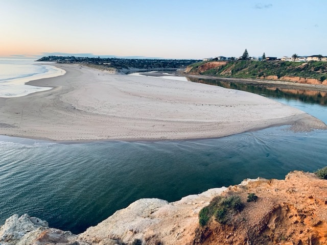 A serene coastal view is captured, showcasing a wide expanse of sandy beach gently curving along the water's edge. The calm river reflects the soft colors of the sky, while distant cliffs and homes can be seen framing the landscape.