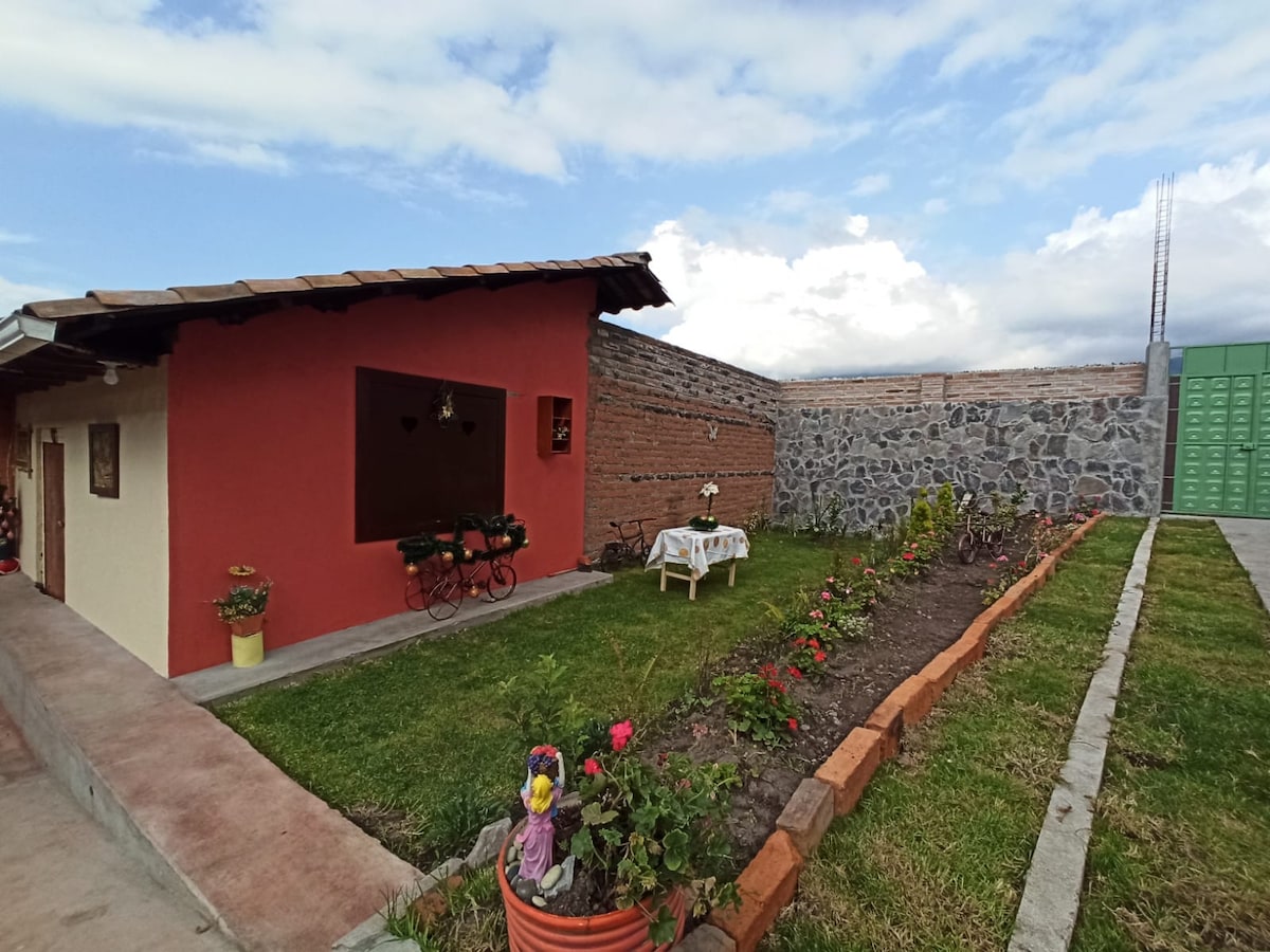 The exterior features a single-story cabin painted in warm colors, with a small garden showcasing neatly arranged flowers and greenery. A table is set in front, accompanied by chairs. A stone wall provides a rustic backdrop, complemented by a clear blue sky.