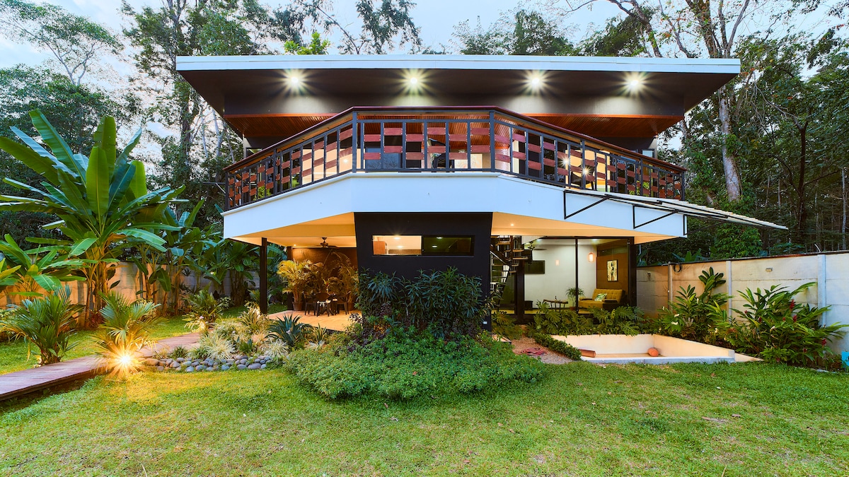 The modern villa is nestled in lush greenery, featuring a unique two-level design. The upper deck showcases an open terrace, while the lower level is framed by vibrant plants and softly lit pathways. Ample windows invite natural light into the space, harmonizing with the surrounding nature.