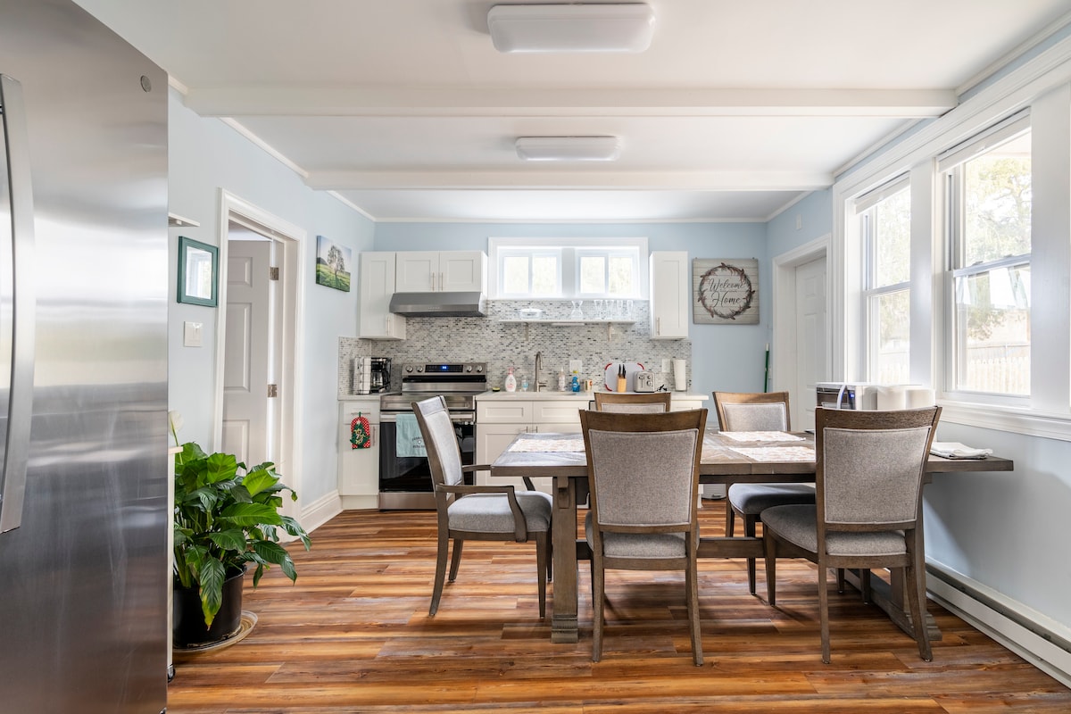 A modern kitchen and dining area is presented, featuring a central wooden table surrounded by six upholstered chairs. Light blue walls complement the stainless steel appliances and white cabinetry. Natural light fills the space through large windows, highlighting the stylish tile backsplash.