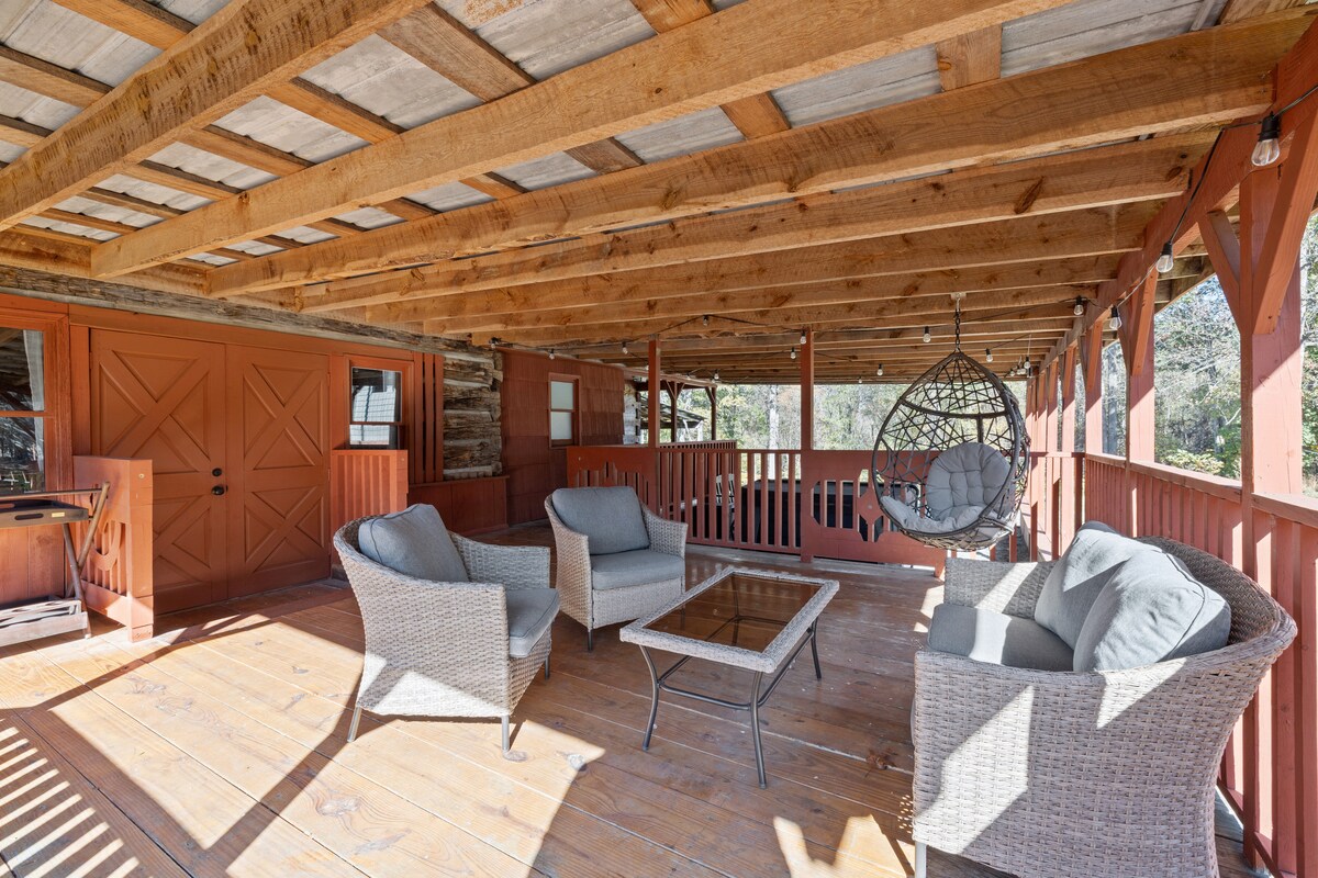 A spacious covered deck features a woven seating arrangement, including four comfortable chairs surrounding a central glass table. A hanging chair adds a relaxed touch, and warm wooden beams support the ceiling, enhancing the natural, inviting setting.