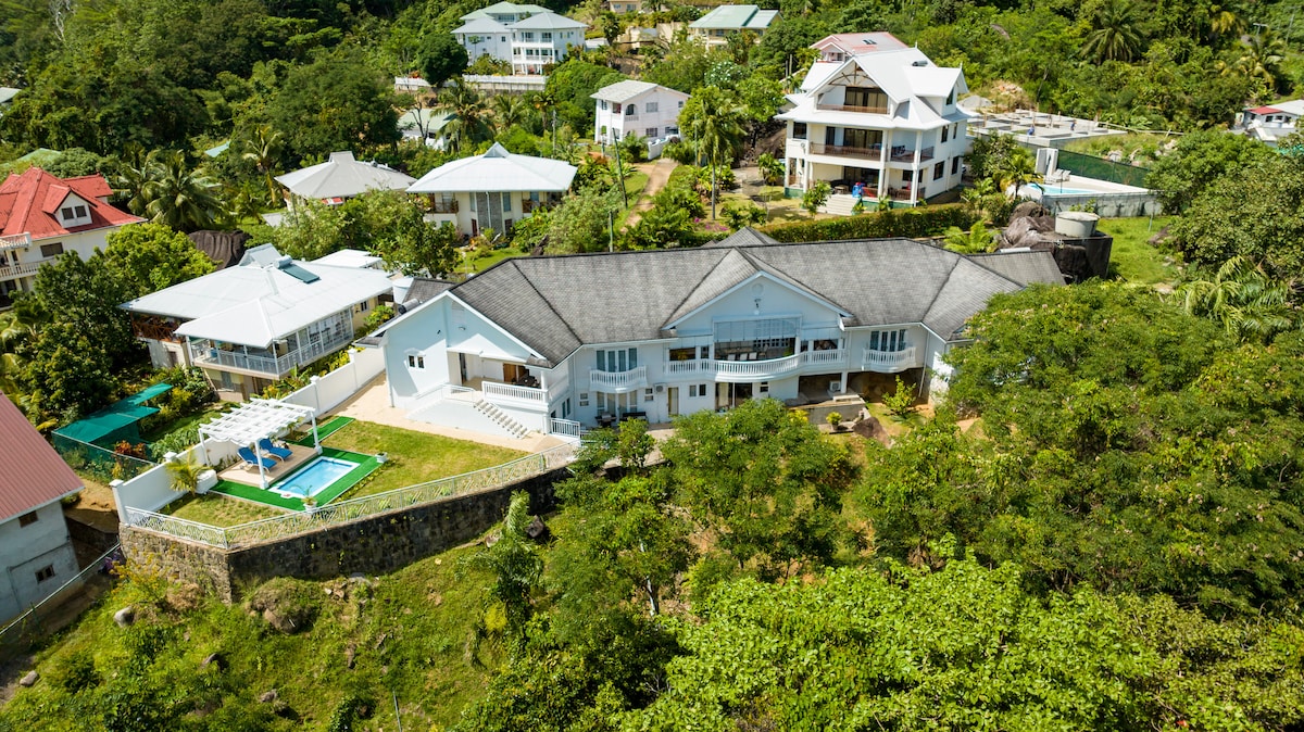 An aerial view of a spacious hillside villa is captured, surrounded by lush greenery. The property features distinct architectural lines, a private pool surrounded by a stone wall, and nearby houses nestled amongst tropical trees, illustrating the tranquil environment of the estate.