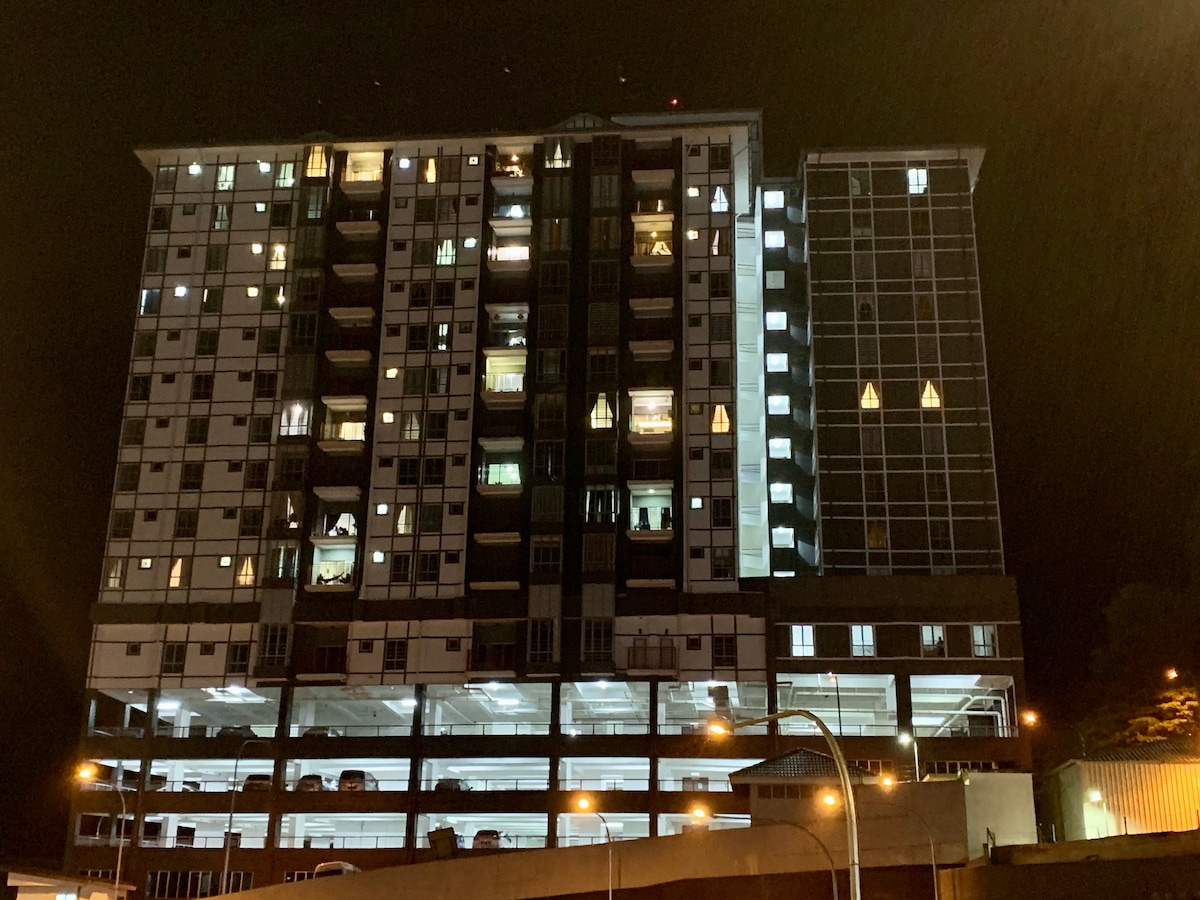 The building is illuminated at night, revealing multiple floors with numerous windows lit from within. The ground level showcases a parking area with several cars visible, while the structure features a mix of dark and light-colored facades.
