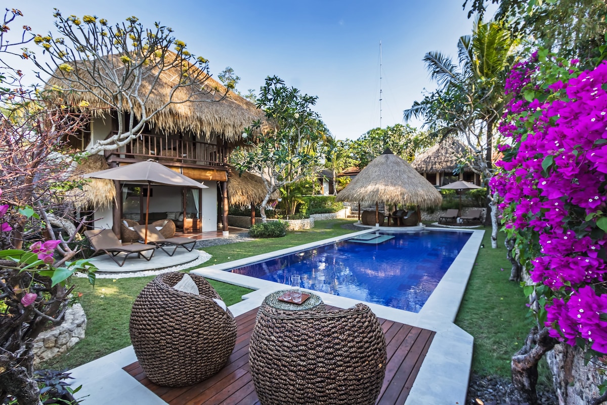 A tranquil garden setting features a central pool surrounded by vibrant tropical flowers and two thatched bungalows. Comfortable seating made of woven material is positioned on a wooden deck, offering a view of the pool and lush greenery.