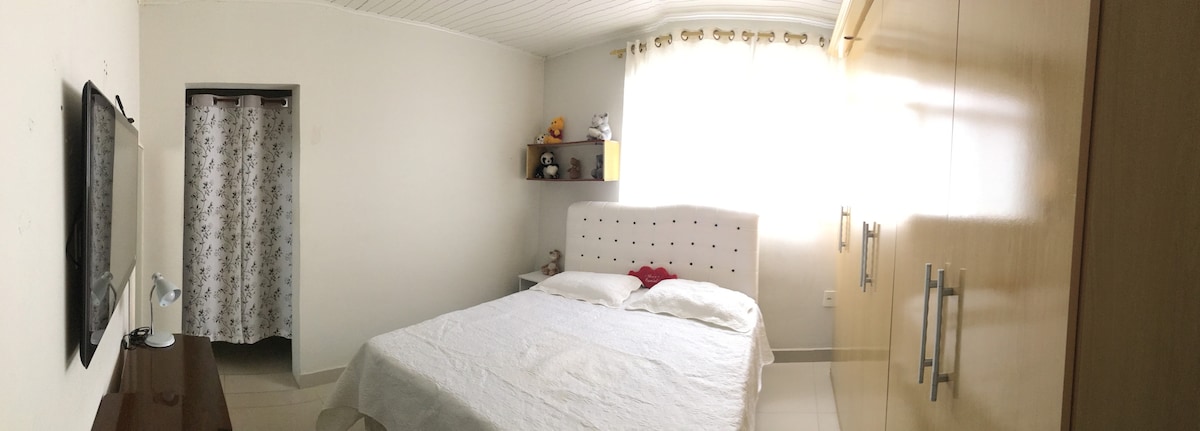 A bright and airy bedroom features a spacious bed with a light-colored coverlet. Soft natural light filters through large curtains, illuminating the room's neutral walls. A wooden dresser sits opposite the bed, with a small bedside lamp and decorative storage above.
