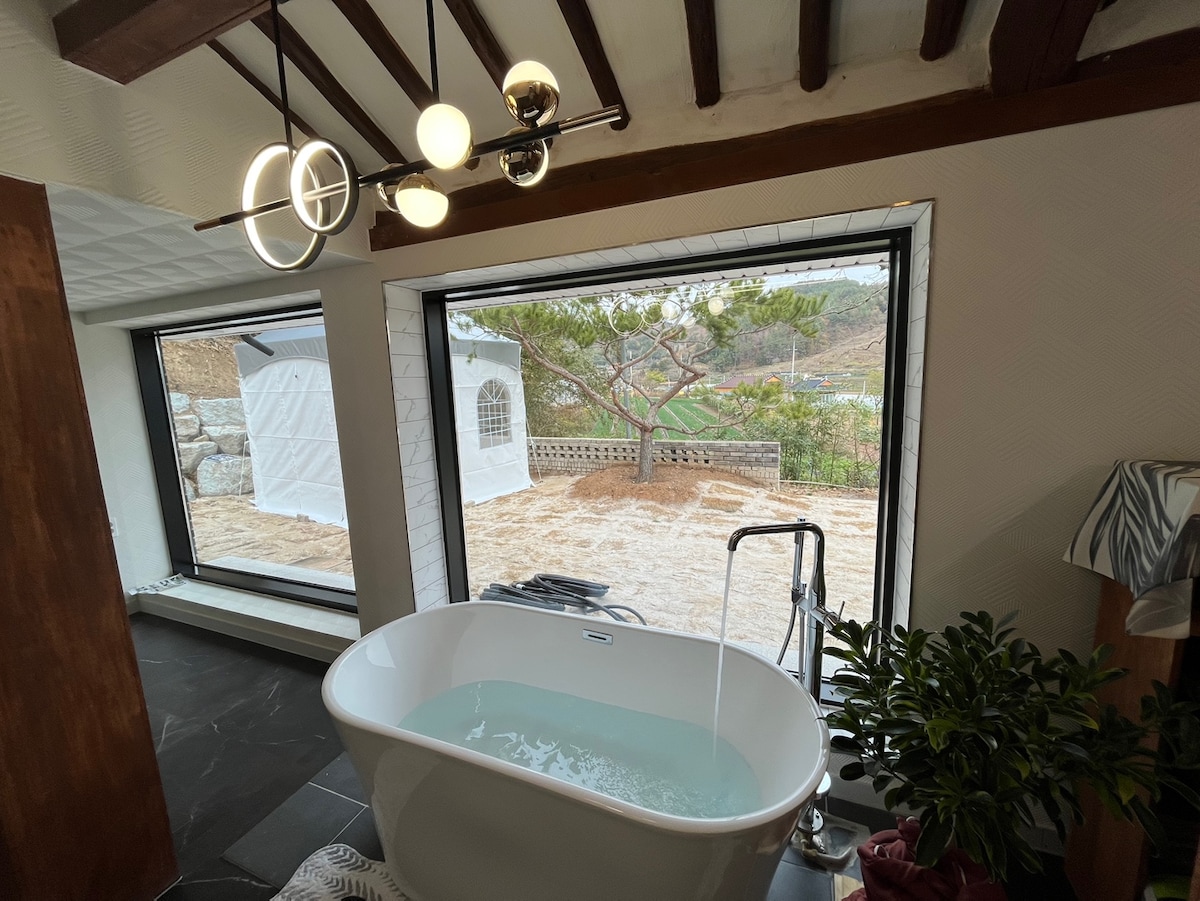A modern freestanding bathtub is featured in the bathroom, positioned beneath large windows that offer a view of the outdoor landscape. Natural light enhances the space, while a potted plant adds a touch of greenery to the interior.
