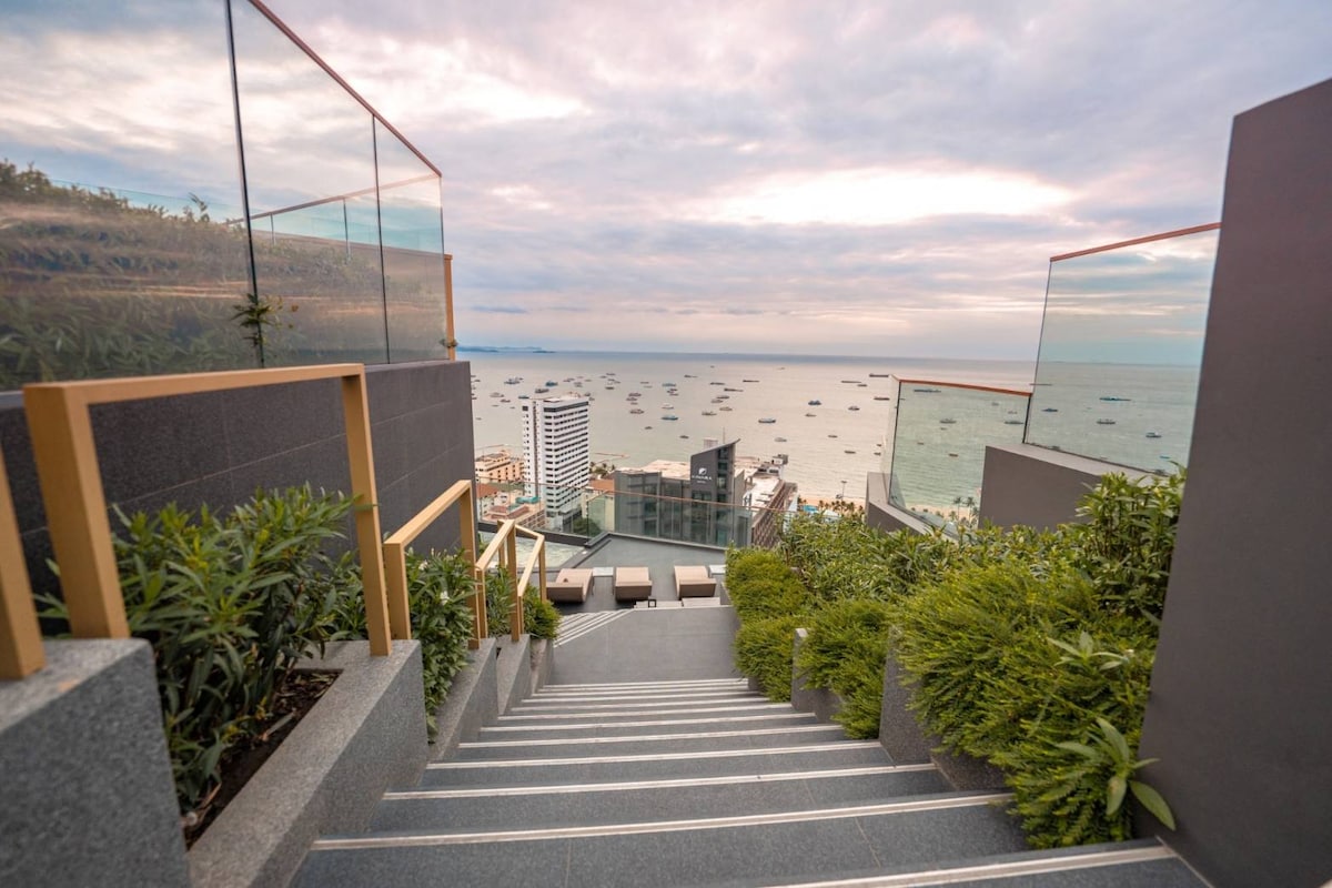 A staircase adorned with lush greenery descends towards a panoramic view of Pattaya Bay. The sky is softly illuminated, with boats dotting the water. The modern high-rise buildings of Pattaya can be seen in the background, providing a vibrant coastal atmosphere.