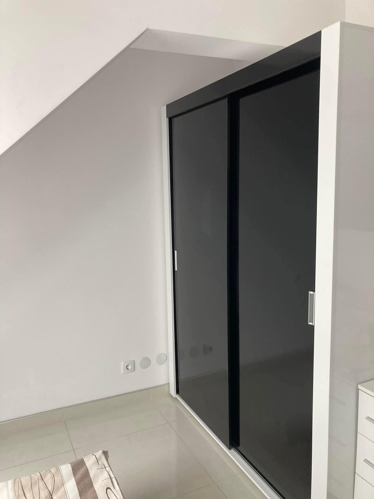 A modern wardrobe features sleek black sliding doors set against a minimalist white wall. The floor is tiled in soft, neutral tones, creating a clean and spacious appearance. Natural light enters from an unseen source, enhancing the clarity of the space.