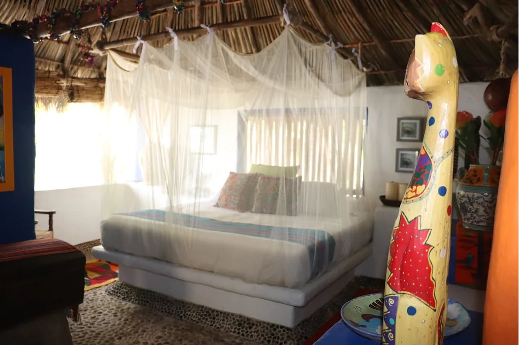 A spacious sleeping area is highlighted by a large bed draped with a lightweight mosquito net, creating a serene atmosphere. Colorful decorative pillows add contrast against the white bedding. Natural light filters through a window, illuminating the room's textured walls and stone flooring.