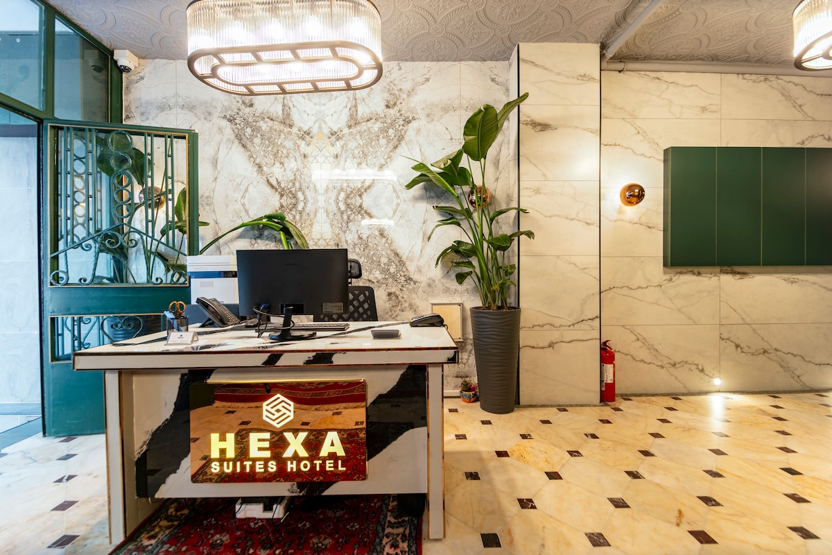 The hotel reception area features a modern design with marble walls and stylish lighting. A large desk with a computer sits at the center, flanked by green plants in decorative pots. An inviting entrance is visible through glass doors, enhancing the open atmosphere.