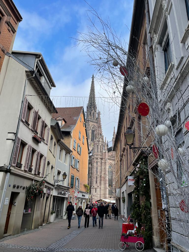 A pedestrian street lined with charming buildings in various colors, leading to a notable church with a tall spire. Seasonal decorations are seen along the street, while pedestrians walk leisurely. A small cart is visible on the right, contributing to the festive scene.