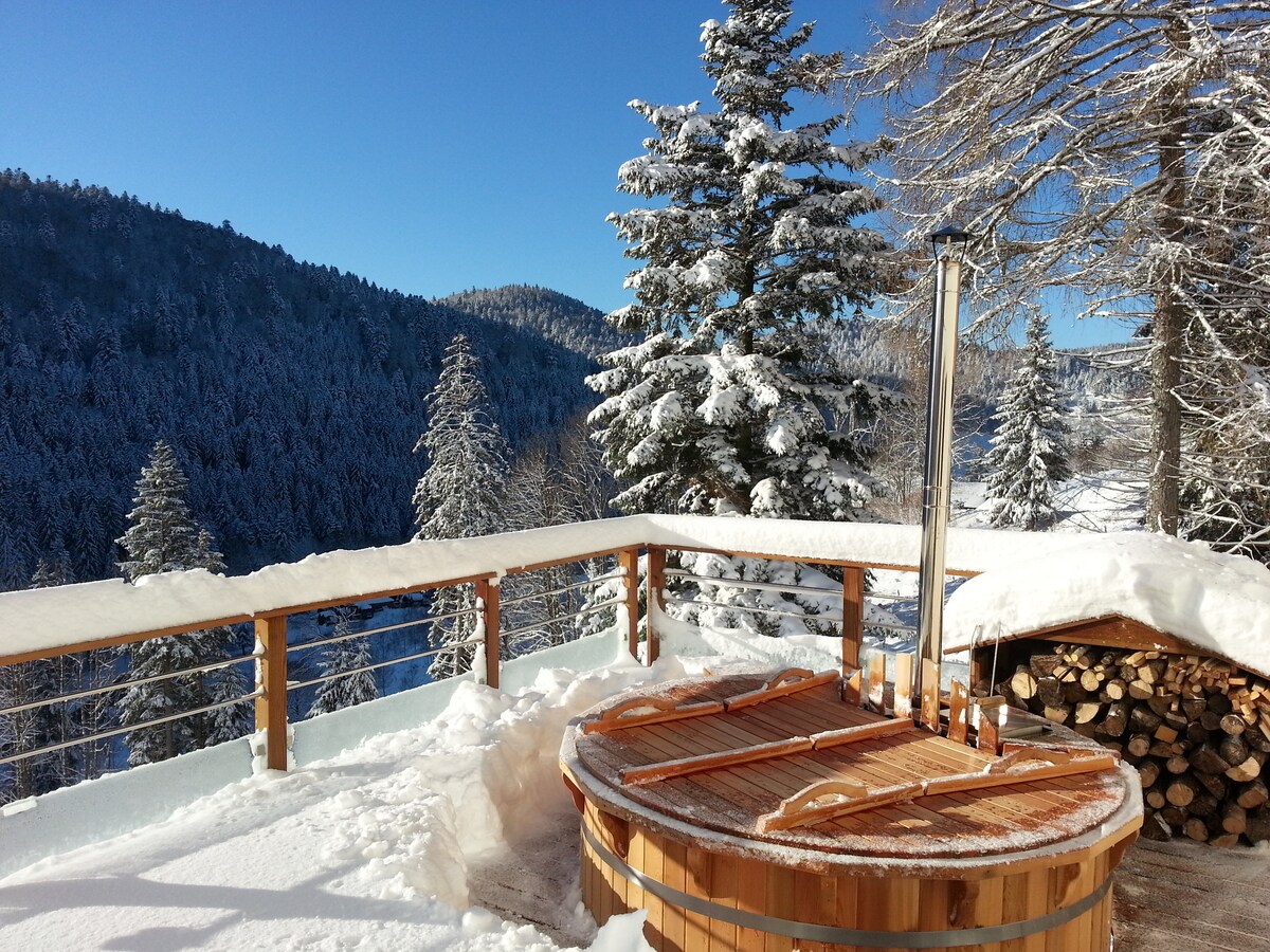 A wooden Scandinavian bath is situated on a snow-covered terrace, surrounded by towering evergreen trees. The landscape features a backdrop of snow-dusted mountains under a clear blue sky, creating a tranquil winter retreat. A stack of firewood is neatly arranged nearby.