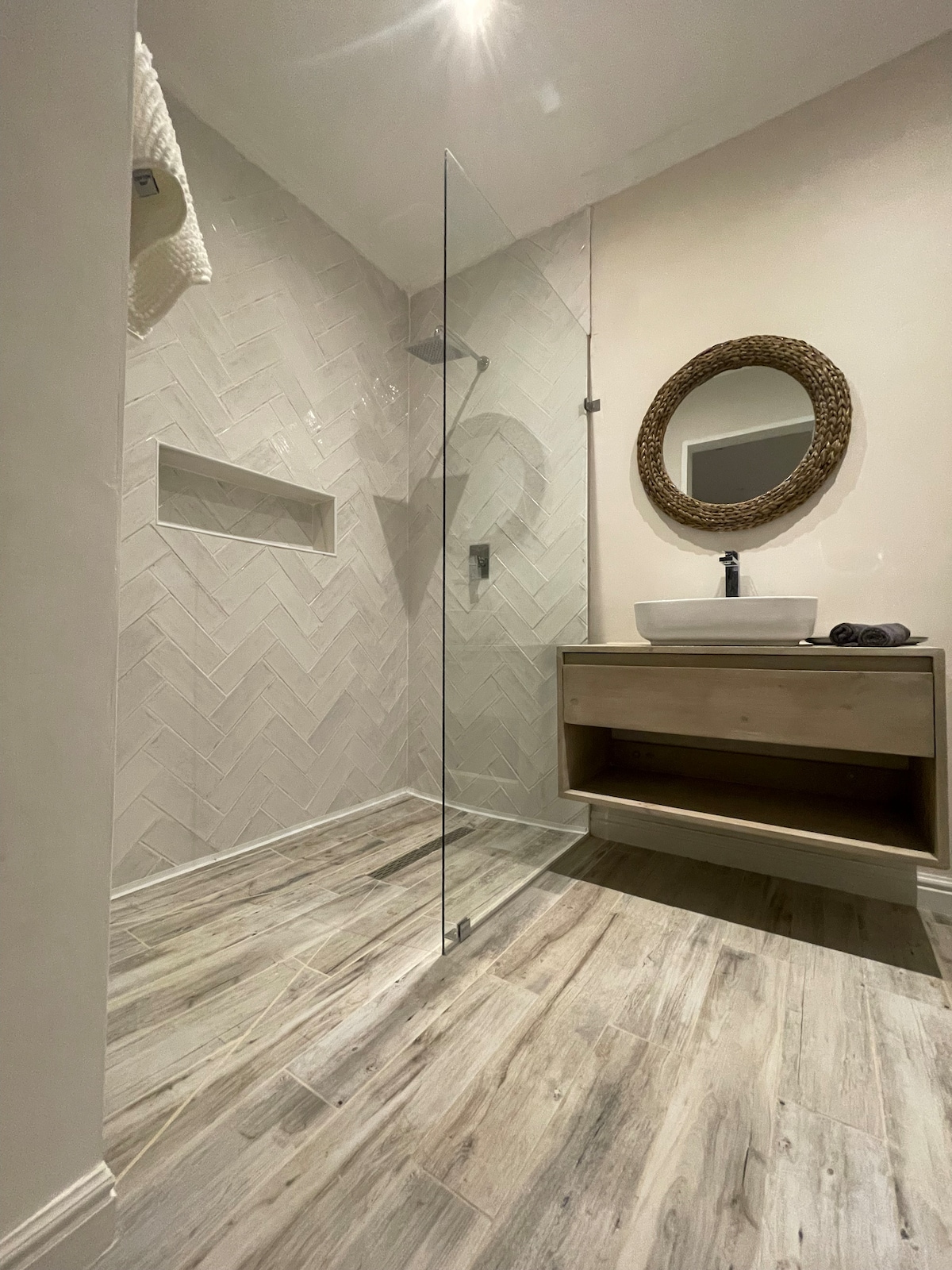 A modern bathroom features a walk-in shower with a glass panel and a rainfall showerhead. A round mirror is mounted above a wooden vanity, complemented by a countertop basin. The herringbone patterned wall tile and wood-look flooring add texture to the space.