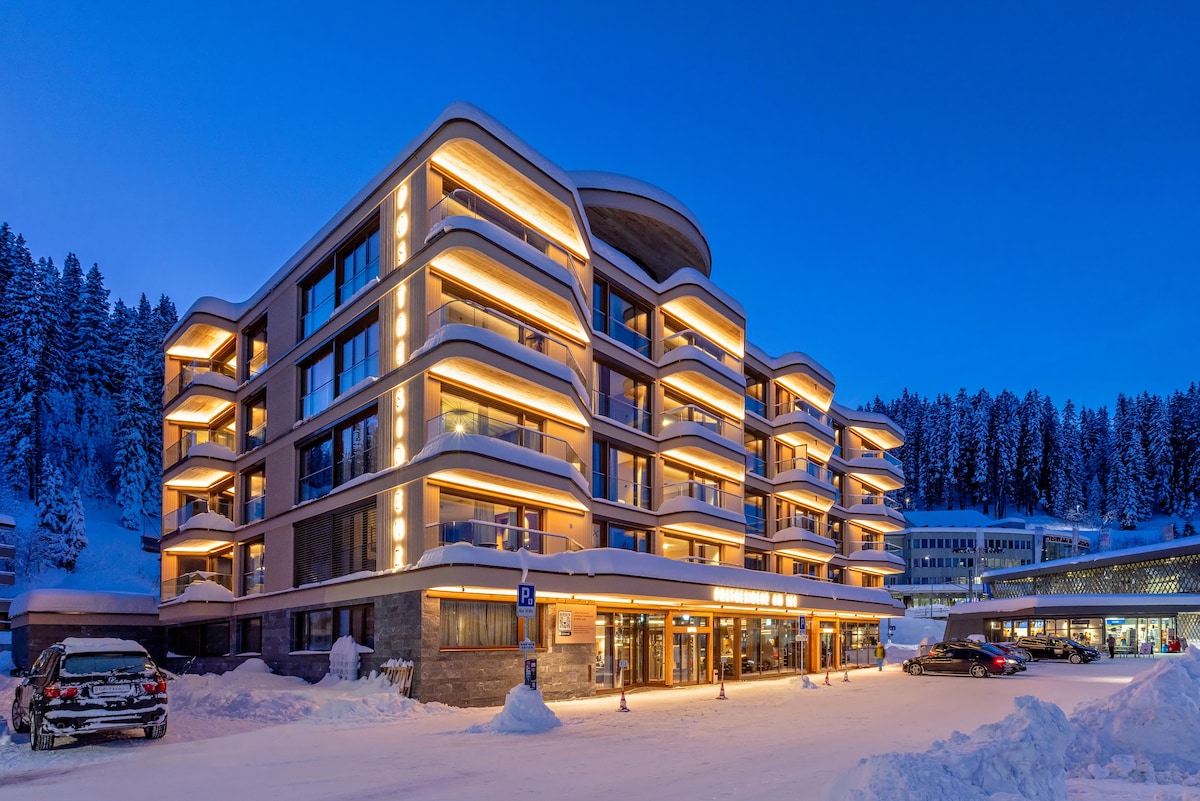 A modern hotel exterior is showcased in a winter setting, featuring soft, warm lighting that highlights architectural details. Snow covers the ground and surrounding trees, creating a serene atmosphere. Multiple balconies are visible, offering an inviting look at the spacious building against a twilight sky.