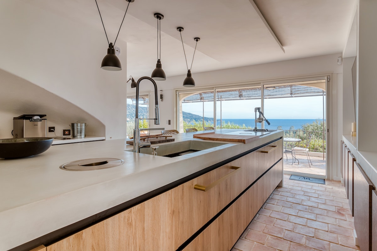 A modern kitchen is featured with wooden cabinetry and an expansive island. Large windows provide a view of the Mediterranean Sea. Pendant lights hang above the kitchen space, and a sleek faucet is positioned near the sink area, enhancing the clean design.
