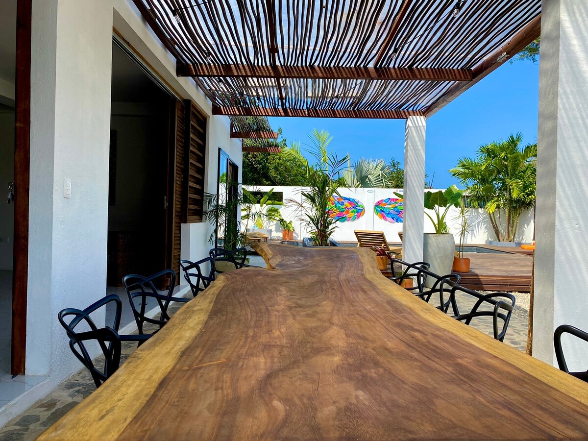An expansive wooden dining table with natural edges is surrounded by black chairs. The space is partially shaded by a wooden lattice, with colorful wall art and lush greenery visible in the background, creating an inviting atmosphere for gatherings.