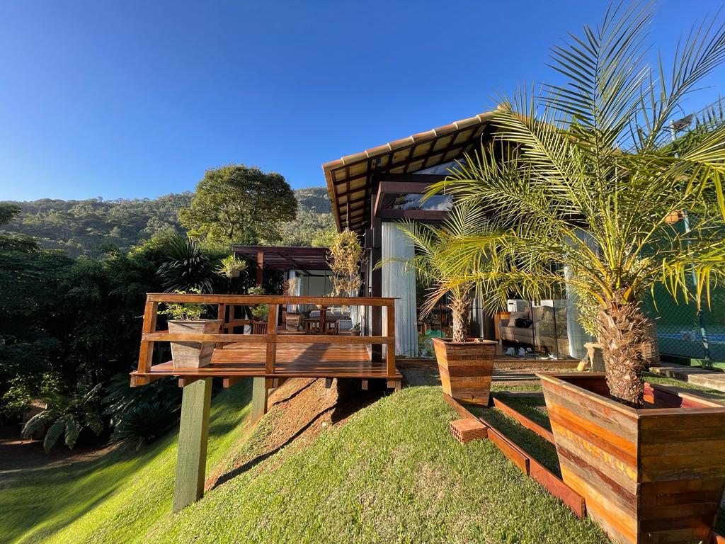 The exterior of the chalet is presented, featuring wooden decks with seating arrangements. Lush greenery surrounds the structure, and tall palm trees are placed in wooden planters. The gentle slope of the landscape leads to nearby mountain views under a clear blue sky.