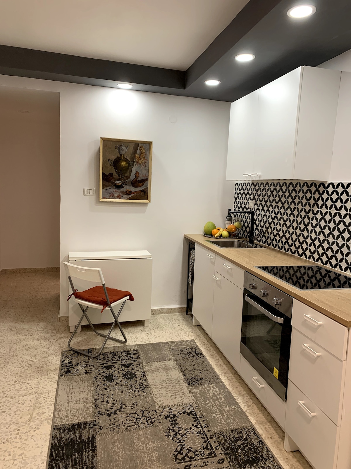 A modern kitchen features white cabinetry and a patterned black and white backsplash. A wooden countertop includes an oven and sink, while a small table with a chair is positioned on a textured rug. Fresh fruits are displayed, adding to the inviting space.