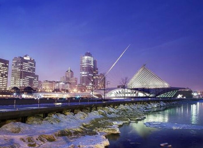 A cityscape at dusk showcases a blend of modern architecture and natural beauty. The shimmering water reflects illuminated buildings, while a striking structure with a unique design stands out against the skyline. A walkway beside the water offers scenic views of the surrounding environment.