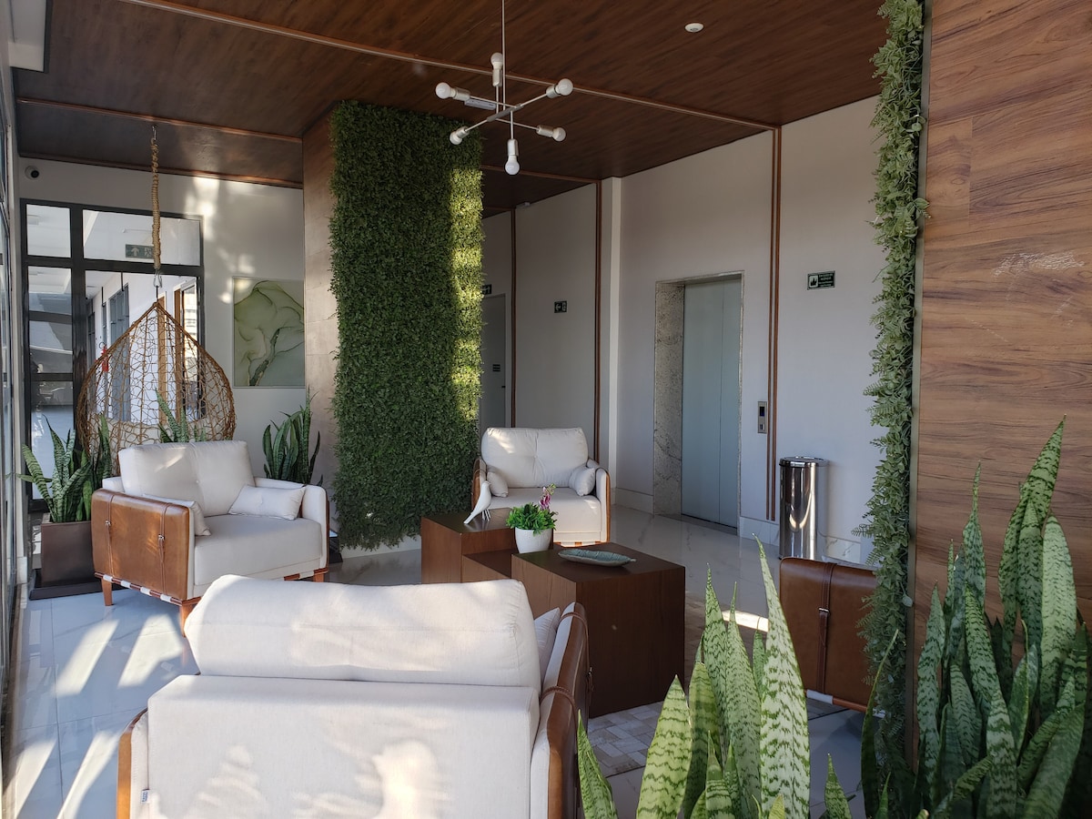 A communal lounge area is featured, with modern comfortable seating arranged around a central table. Lush green plants add a natural touch, and light enters through large windows, creating a bright environment. Elevator access is visible in the background.