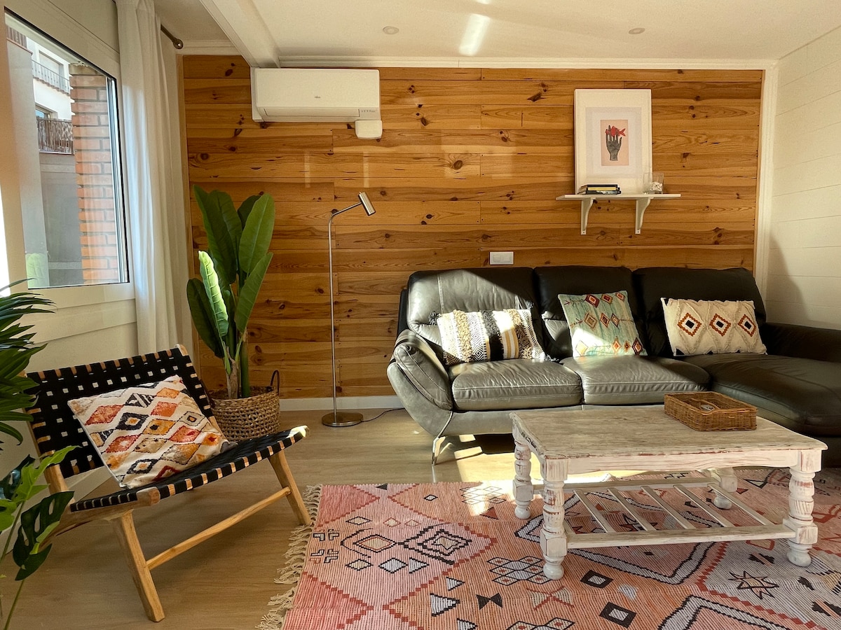 A comfortable living area features a leather sofa with decorative cushions, complemented by a coffee table and a cozy patterned rug. A floor lamp and a potted plant add warmth, while light streams in through large windows, creating a bright and inviting ambiance.