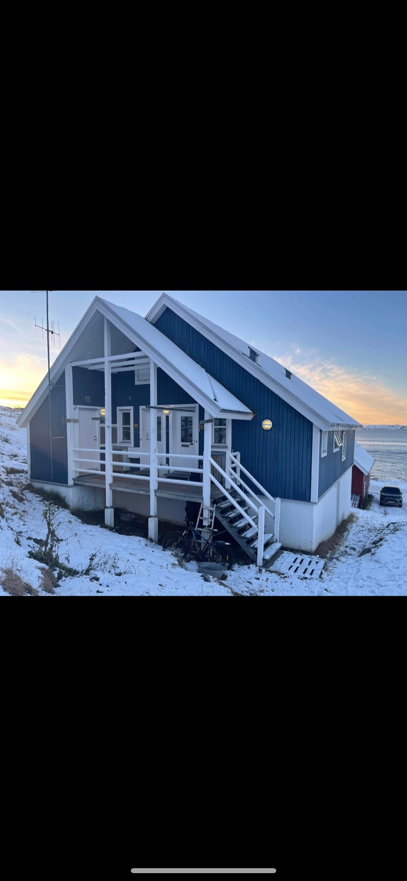 A charming A-frame cabin features a blue exterior with white trimmings and multiple large windows. A staircase leads to the entrance, while snow blankets the ground surrounding the home. The peaceful landscape includes a distant coastline visible under a soft sunrise.