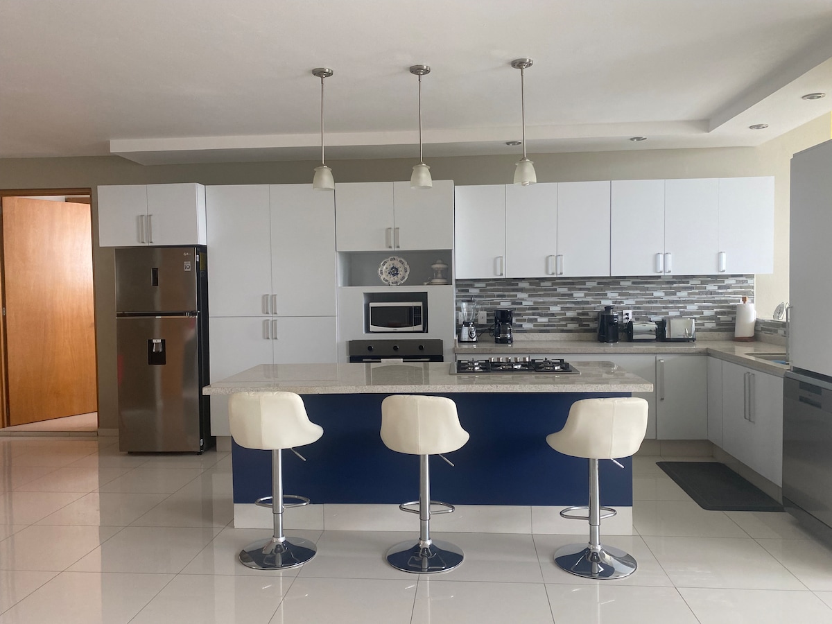 The modern kitchen features a central island with three bar stools and a gas cooktop. White cabinetry is complemented by stainless steel appliances and a mosaic backsplash. Large windows allow natural light to illuminate the spacious area, enhancing its clean and functional design.
