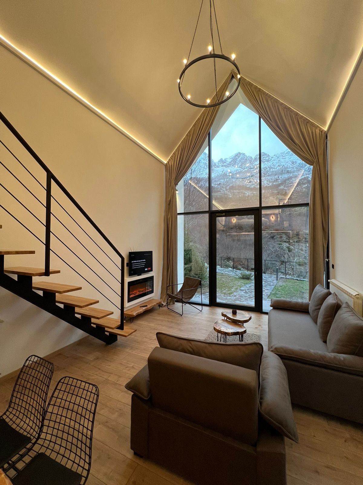 A modern living area features a spacious sofa and a sleek TV stand. Large glass doors provide a view of the mountainous landscape, while a warm ambiance is enhanced by soft lighting. A staircase leads to the upper level, adding an element of depth to the design.