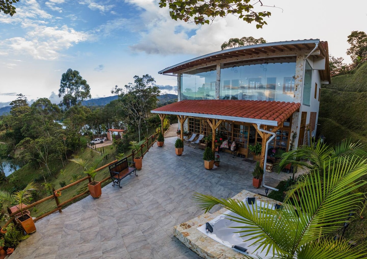 The spacious terrace of the Finca is surrounded by lush greenery and features a large glass front that offers expansive views of the landscape. Several lounge chairs are positioned for relaxation, and a Jacuzzi awaits for a soothing experience.