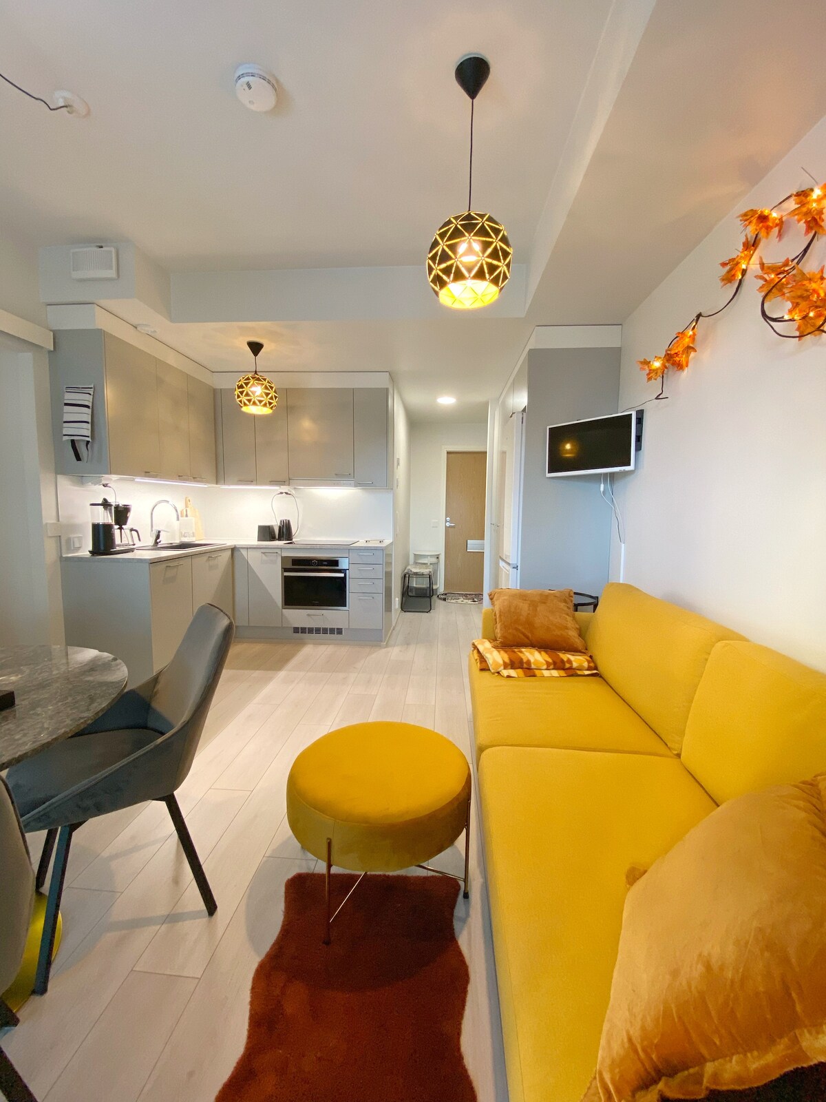 A modern living space features a yellow sofa complemented by a small round ottoman. The kitchen area showcases sleek cabinetry and appliances, while a dining table is visible. Warm pendant lights hang from the ceiling, adding a cozy touch to the room.