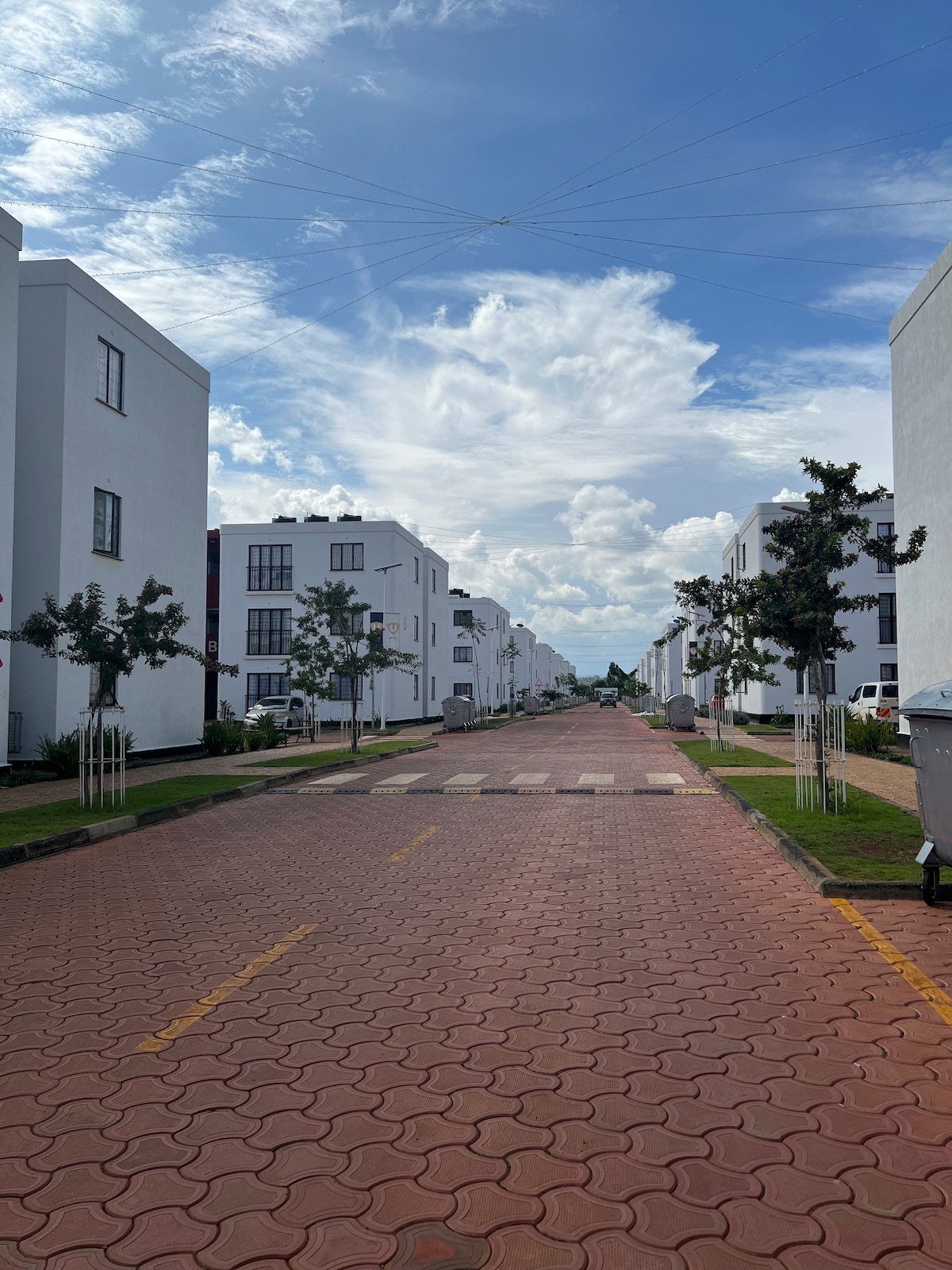 A tree-lined street is flanked by modern, white multi-story buildings featuring large windows. Fluffy clouds drift across a blue sky, providing a serene backdrop. The wide pathway is paved with red bricks, and greenery can be seen along the edges.
