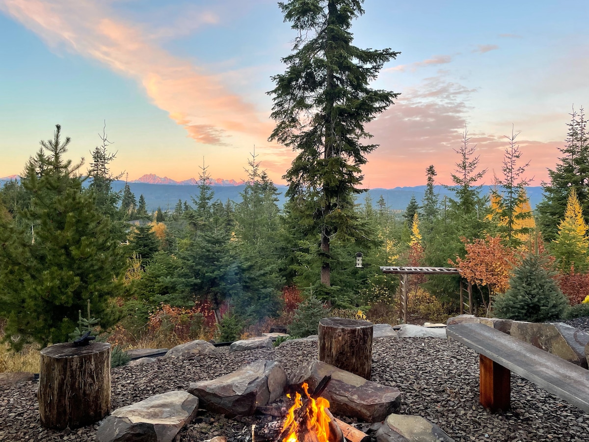 A stone firepit sits surrounded by log seating, providing a cozy gathering space. Towering trees create a natural backdrop, while distant mountain views are visible under a pastel sky at dusk. The warm glow from the fire contrasts with the cool colors of the landscape.