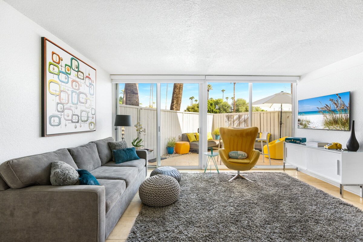 A light-filled living space features a comfortable gray sofa adorned with decorative pillows. A vibrant yellow accent chair is positioned near sliding glass doors that open to a private outdoor patio, showcasing palm trees and outdoor seating. A cozy ottoman is placed on a plush area rug.