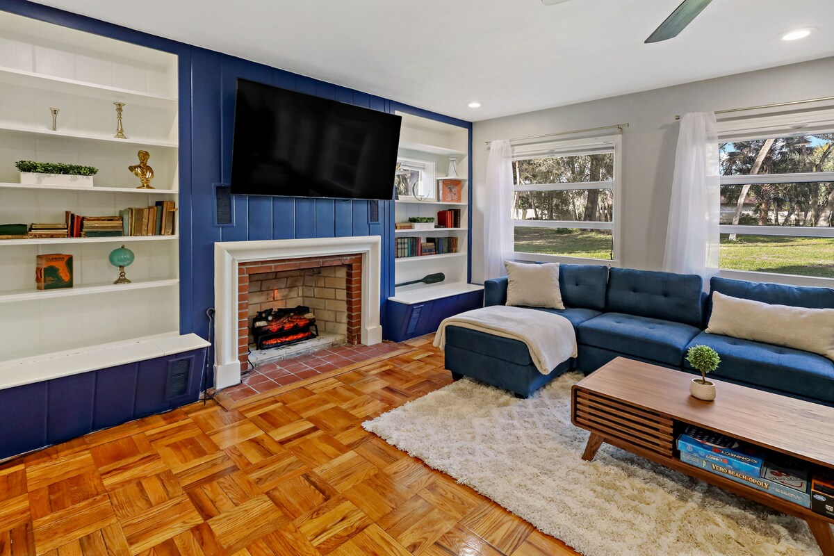 The living room features a stylish sectional sofa positioned near a modern coffee table. A wall-mounted smart TV is above a traditional fireplace. Natural light floods the space through large windows, illuminating the inviting hardwood floor.