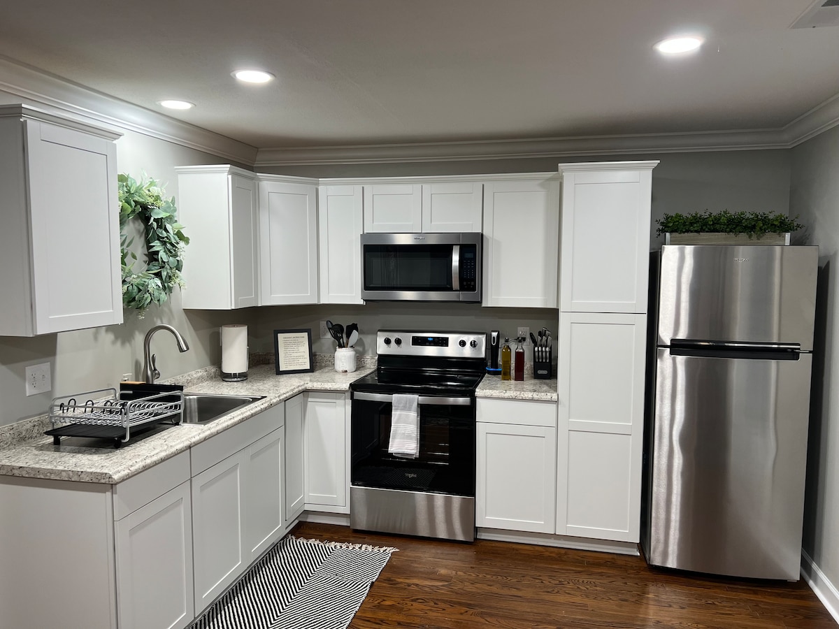 The kitchen features modern white cabinetry complemented by granite countertops. Stainless steel appliances, including a microwave and refrigerator, are visible. A dish rack is positioned near the sink, and a striped rug lies on the floor, adding a touch of texture to the space.