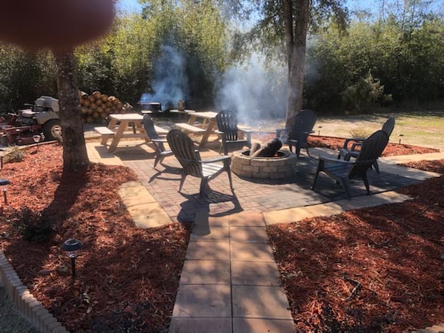 An outdoor fire pit area reveals multiple adirondack chairs arranged around a stone circle. A wooden picnic table sits nearby, surrounded by fresh landscaping and mulch, creating a comfortable gathering space. Smoke rises from the fire, while greenery provides a natural backdrop.