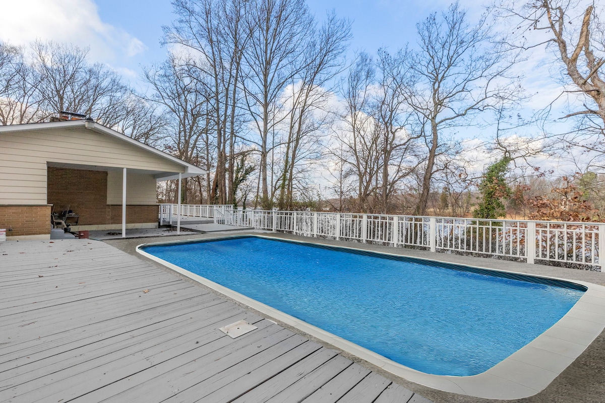 A private outdoor deck features a large swimming pool surrounded by white railings. The area is partially shaded by trees, with a view of the water in the background. The deck provides space for lounging, making it ideal for relaxation and enjoyment.