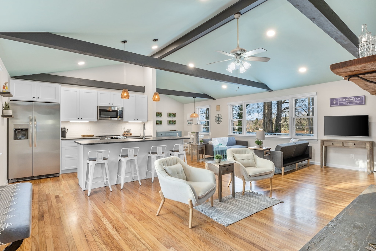 An open floor plan features a modern kitchen with stainless steel appliances and a spacious island. Comfortable seating arrangements are visible on polished hardwood floors, under high ceilings with exposed beams. Large windows offer generous natural light and views of the surrounding landscape.