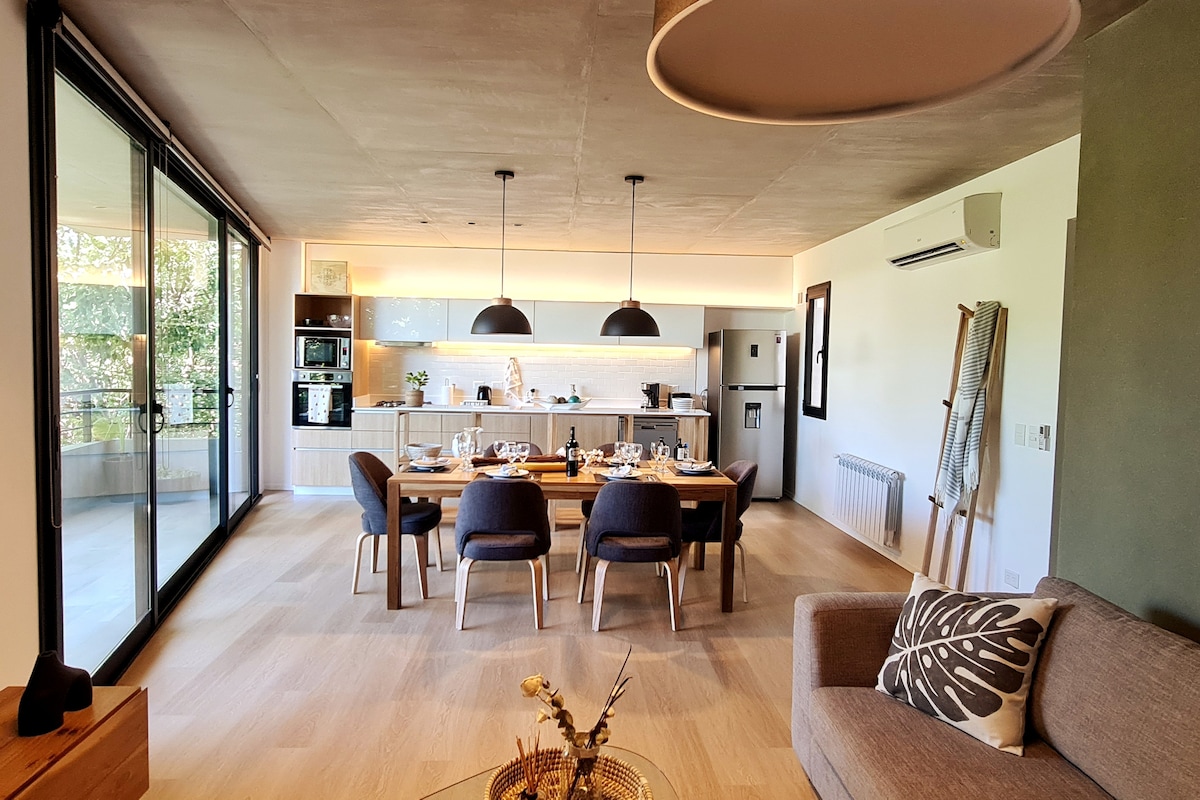 An open living area presents a dining table set for six, surrounded by wooden chairs. A modern kitchen, equipped with sleek appliances, is visible in the background. Large glass doors allow natural light to fill the space, while offering a view of an outdoor balcony.