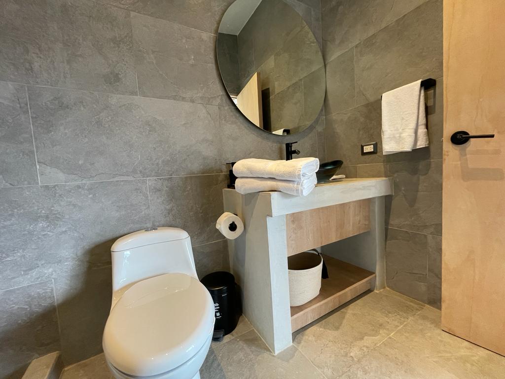 A well-appointed bathroom features a modern toilet and a stylish vanity with a round mirror above it. Soft, folded towels are neatly arranged, and a roll of toilet paper is visible. The space is accented by natural stone tiles, creating a calming atmosphere.