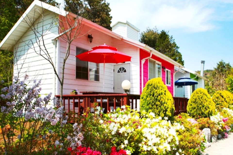 A charming single-story house is framed by vibrant flower beds and manicured shrubs. The exterior features a mix of white and pink siding, with red and blue umbrellas placed on the front deck, creating a welcoming outdoor space for relaxation.