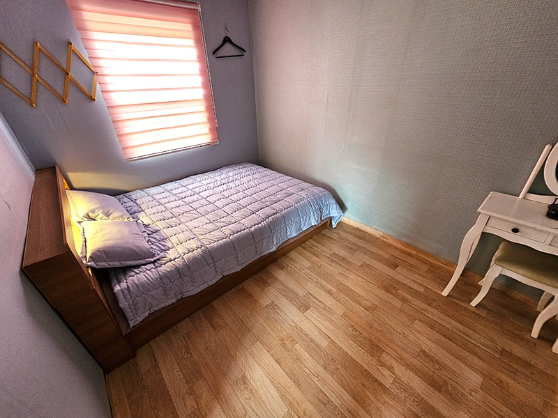 This room features a spacious bed with a light quilt and a simple wooden headboard. A small white vanity table sits against the wall, and a window with layered shades provides soft, diffused light in the space.