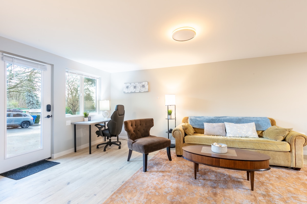 A living room area is depicted with a comfortable sofa and an accent chair positioned around a coffee table. Natural light enters through a large window. A desk with an office chair is visible, providing a workspace option. A light fixture illuminates the space.