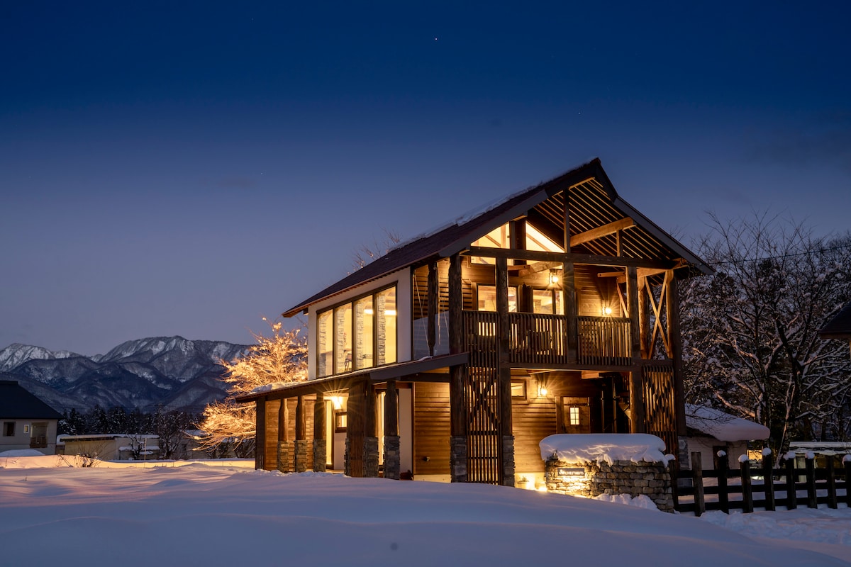 A two-story cottage is illuminated by soft lighting against the twilight sky. Snow blankets the ground, while the surrounding mountains provide a serene backdrop. The design features large windows that enhance the connection with the outdoors.