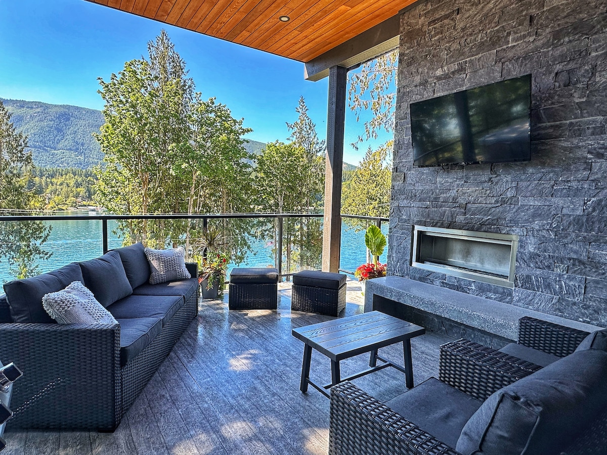 An outdoor living space is showcased with comfortable seating arranged around a table, featuring a fireplace built into the stone wall. Lush greenery and lake views create a serene atmosphere, while a wall-mounted television adds modern convenience.