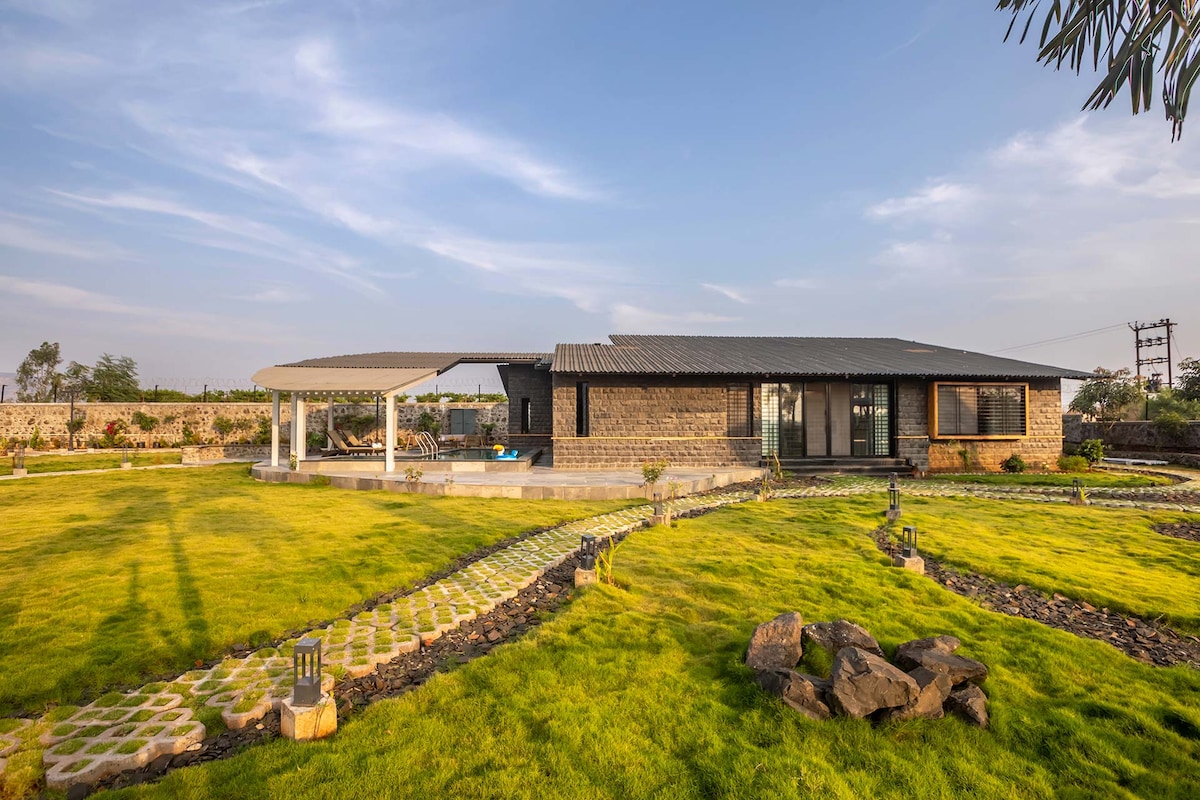 The spacious holiday home is surrounded by lush green lawns, with a stone pathway leading up to the inviting entrance. A shaded patio area provides outdoor seating, and a private pool is positioned to the side, offering a tranquil retreat amidst the natural landscape.