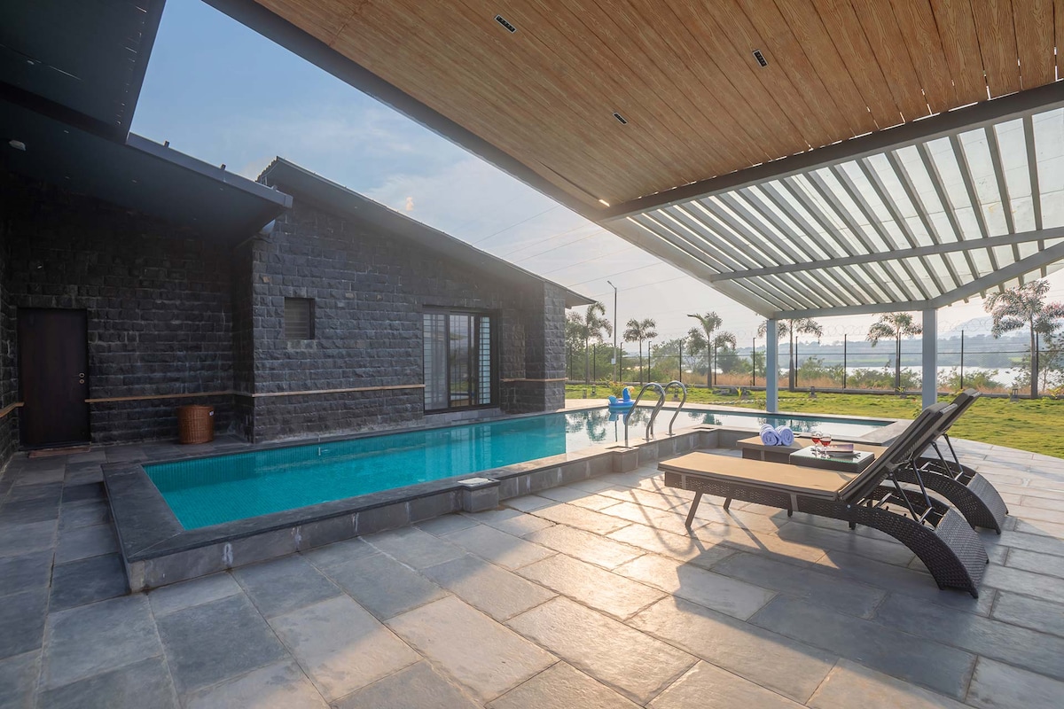 A private swimming pool is bordered by a spacious deck featuring lounge chairs. The pool area is shaded by a roof, allowing for comfortable relaxation. Lush greenery is visible in the background, alongside a scenic view of the dam.