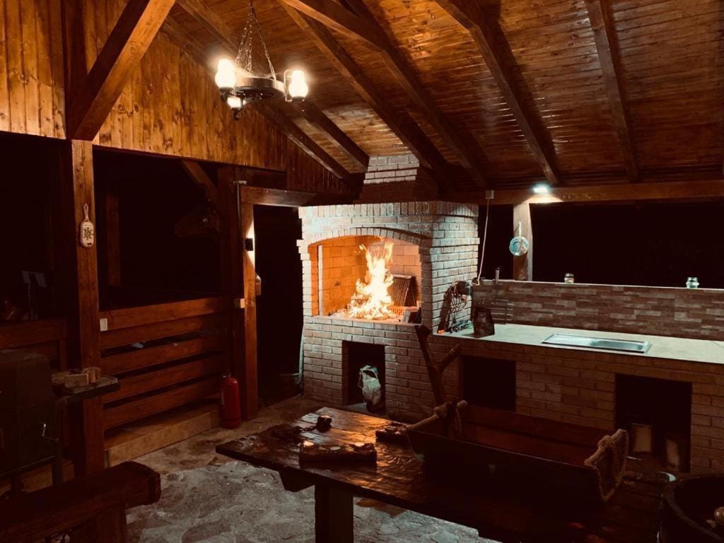 An outdoor kitchen area features a brick fireplace with a warm fire crackling inside. Wooden beams and a high ceiling create an inviting space. A stone table is positioned nearby, with benches arranged around it, enhancing the atmosphere for gatherings. Subtle lighting adds to the overall ambiance.